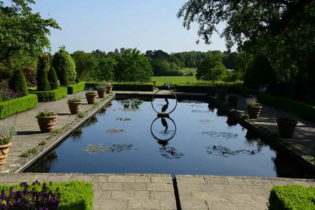 Ein rechteckiger, spiegelnder Teich mit Topfpflanzen säumt einen steinernen Weg, der von grünen Bäumen und Sträuchern umgeben ist und in dessen Mitte eine kreisförmige Skulptur mit einem Vogel steht - eine Reminiszenz an die Chelsea Flower Show unter strahlendem, sonnigem Himmel.