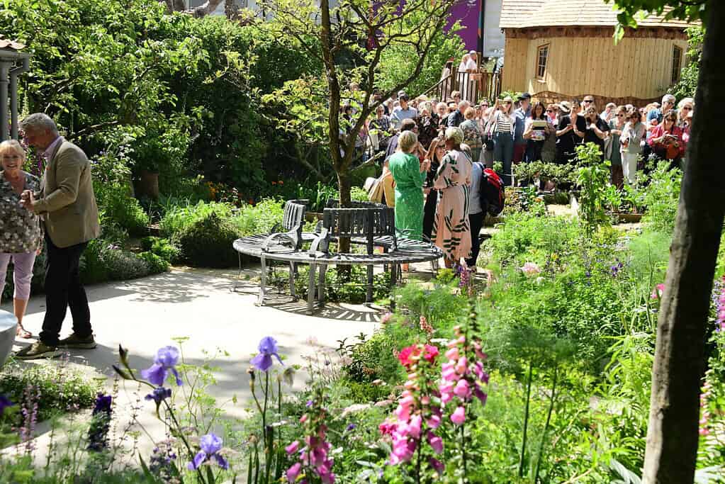 Ein üppiger Garten mit blühenden Blumen und Grünzeug umgibt eine runde Bank. Im Hintergrund sind Menschen versammelt, die sich mischen und spazieren gehen und eine sonnige Veranstaltung im Freien genießen, die an die Chelsea Flower Show in der Nähe eines Holzgebäudes erinnert.