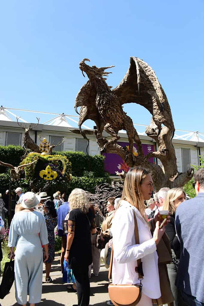 Eine Menschenmenge versammelt sich bei der Chelsea Flower Show unter freiem Himmel vor einer großen, drachenförmigen Holzskulptur mit ausgebreiteten Flügeln.