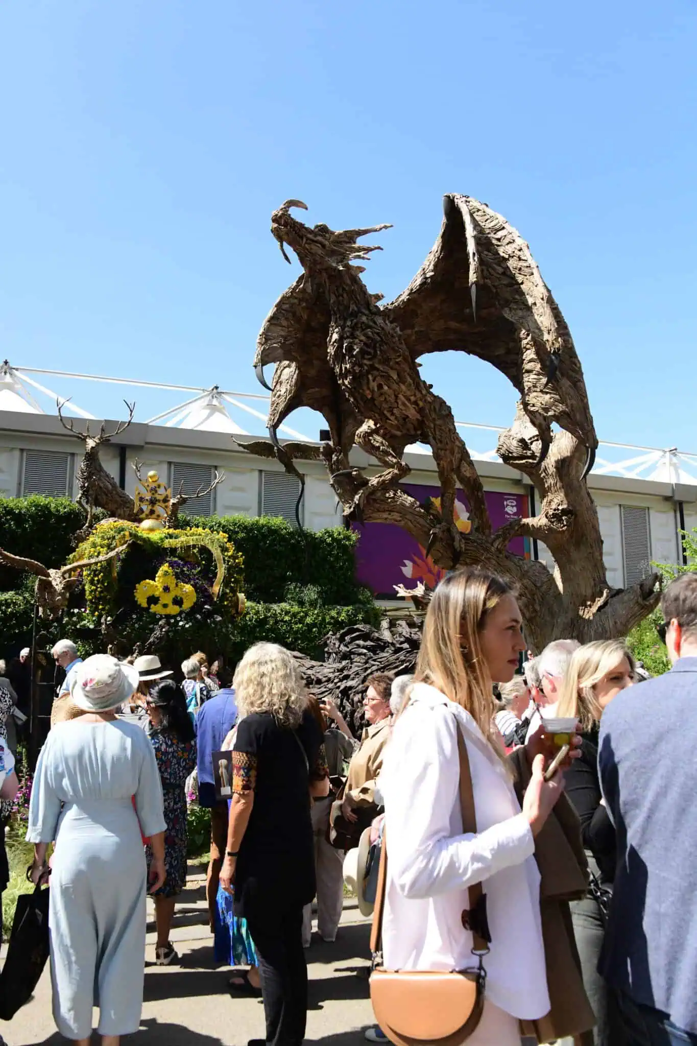 Eine Menschenmenge versammelt sich bei der Chelsea Flower Show unter freiem Himmel vor einer großen, drachenförmigen Holzskulptur mit ausgebreiteten Flügeln.