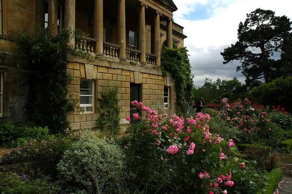 Ein historisches Steingebäude mit Säulen blickt auf einen üppigen Garten voller blühender rosa Rosen, grüner Sträucher und hoher Bäume unter einem teilweise bewölkten Himmel.