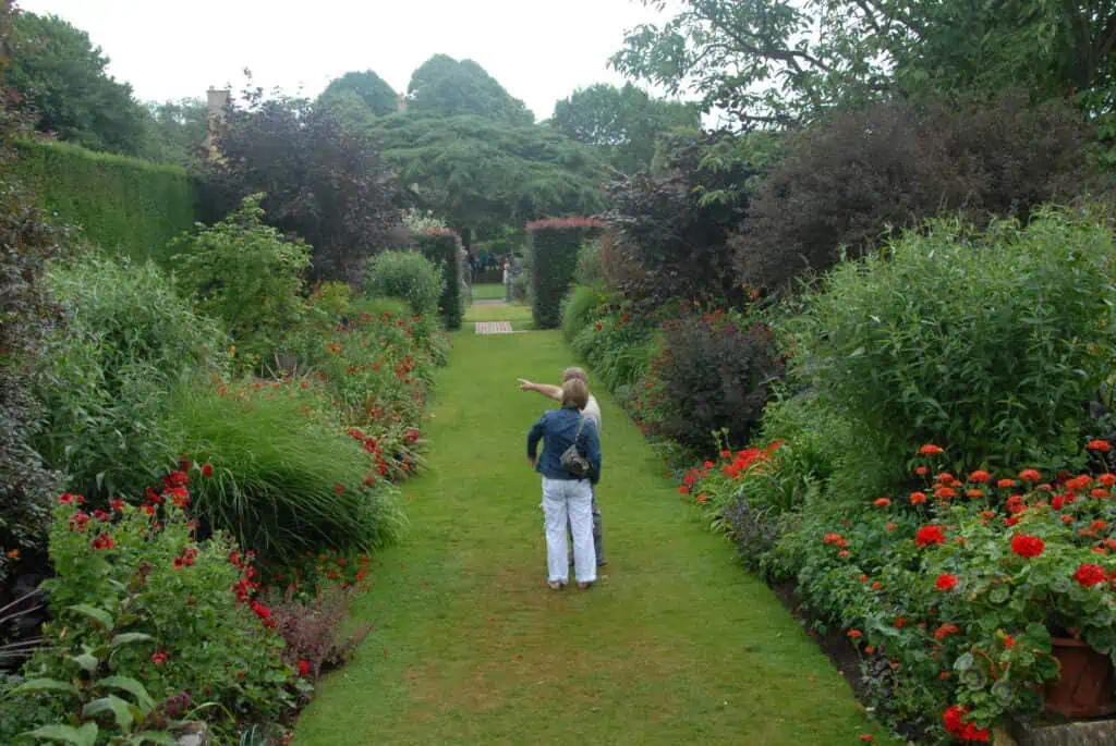 Zwei Personen stehen auf einem grasbewachsenen Weg in einem Garten, der von bunten Blumen und üppigen grünen Büschen gesäumt ist. Eine Person zeigt nach vorne, während die andere daneben steht, beide mit dem Blick von der Kamera weg.