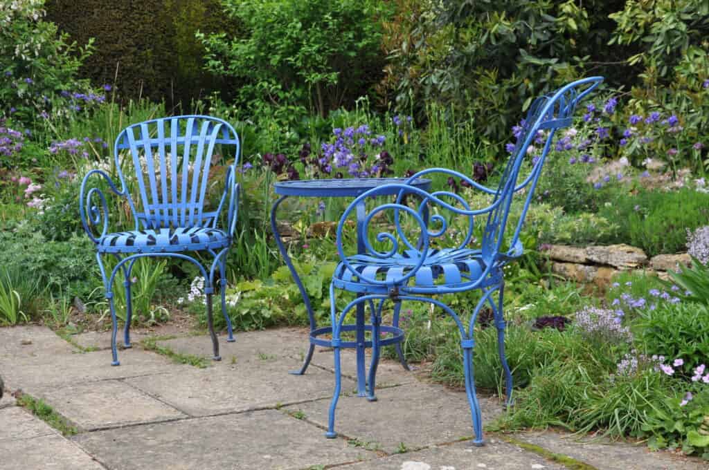 Zwei blaue Metallstühle und ein passender runder Tisch stehen auf einer steinernen Terrasse, umgeben von üppigen grünen Pflanzen und lila Blumen in einer Gartenlandschaft.