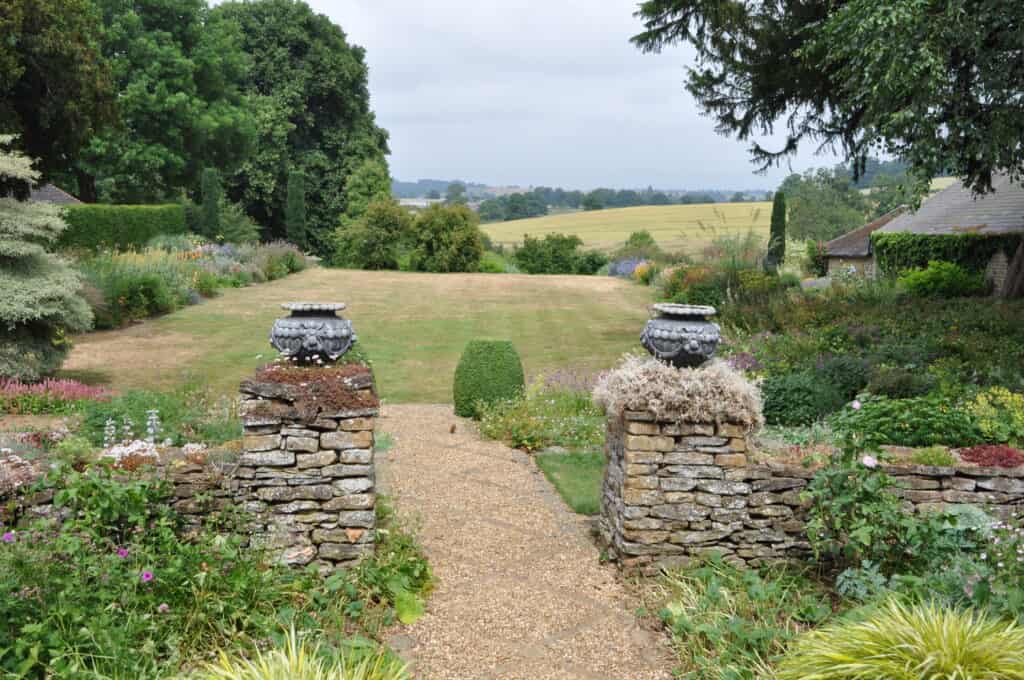 Ein Kiesweg führt durch einen Landschaftsgarten mit Steinmauern und Urnen und öffnet sich zu einer großen Rasenfläche, die von bunten Blumen und Grünpflanzen gesäumt ist, mit Feldern und Bäumen in der Ferne unter einem bewölkten Himmel.