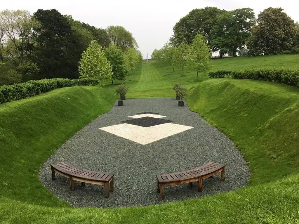Zwei geschwungene Holzbänke stehen vor einer geometrischen Kiesfläche mit schwarzen und weißen Formen, umgeben von abfallenden Grasflächen und üppig grünen Bäumen in einer Parklandschaft.