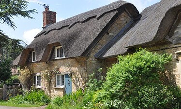 Ein charmantes Steinhaus mit Strohdach, weiß gerahmten Fenstern und einem gemauerten Schornstein, umgeben von üppigem Grün und Pflanzen an einem sonnigen Tag.