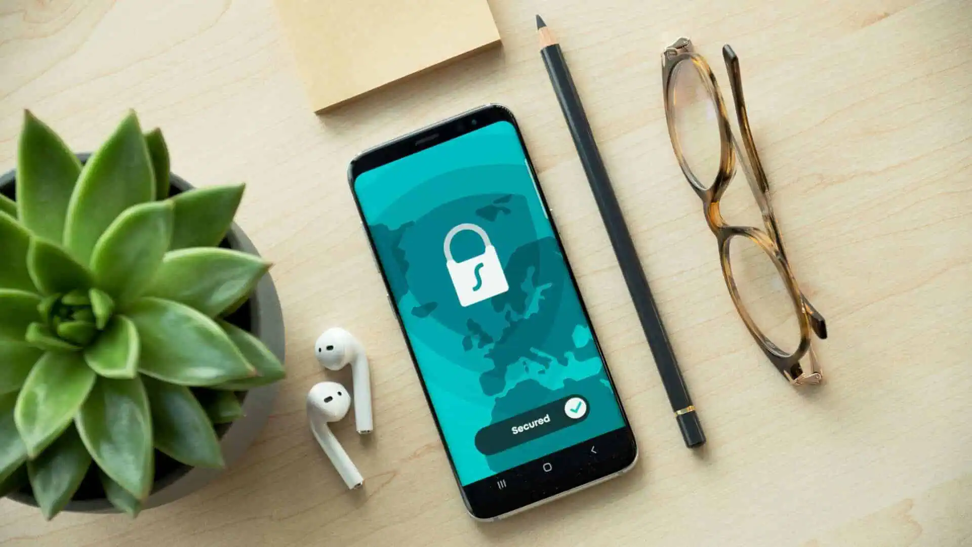 Ein Smartphone mit einem Sicherheitsschloss-Symbol, das Datenschutz und DSGVO symbolisiert, liegt auf einem Schreibtisch neben einer eingetopften Sukkulente, einer Brille, einem Bleistift, einem Haftnotizblock und weißen kabellosen Ohrhörern.