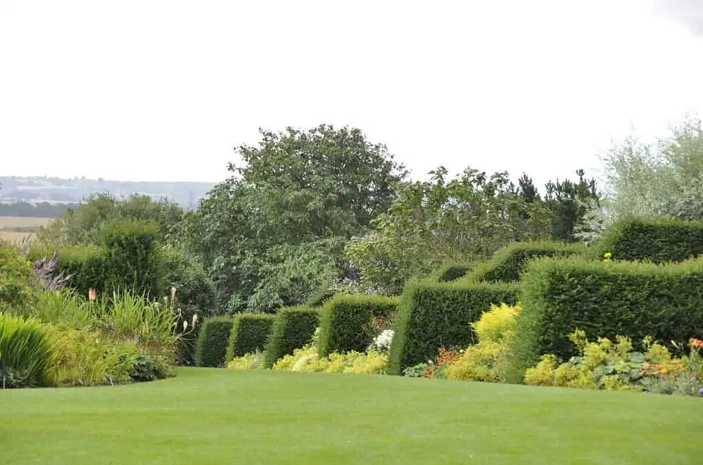 Ein gepflegter Garten mit ordentlich geschnittenen, quadratischen Hecken, farbenfrohen Blumenbeeten und saftig grünem Rasen. Im Hintergrund sieht man Bäume und offene Landschaften unter einem bewölkten Himmel.