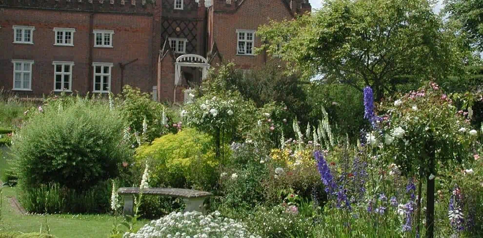 Ein historisches Backsteinhaus mit vielen hohen Schornsteinen steht hinter einem üppigen, farbenfrohen Garten mit blühenden Pflanzen, Sträuchern und einer Steinbank unter einem bewölkten Himmel.
