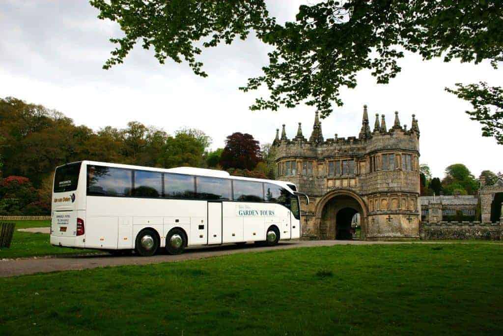 Ein weißer Reisebus mit der Aufschrift Garden Tours wartet an einer der Einstiegsstellen auf einem Weg vor einem historischen Steingebäude mit einem gewölbten Eingang, umgeben von grünem Gras und Bäumen.