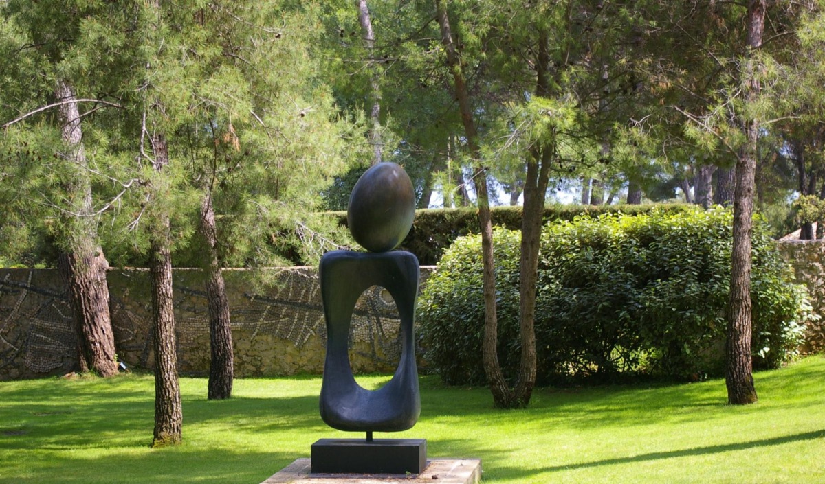 Eine moderne abstrakte Skulptur mit einer großen ovalen Form, die auf einem hohlen, geschwungenen Sockel balanciert, steht auf einem Sockel in einem sonnenbeschienenen Garten, der von Bäumen und Grünpflanzen umgeben ist.