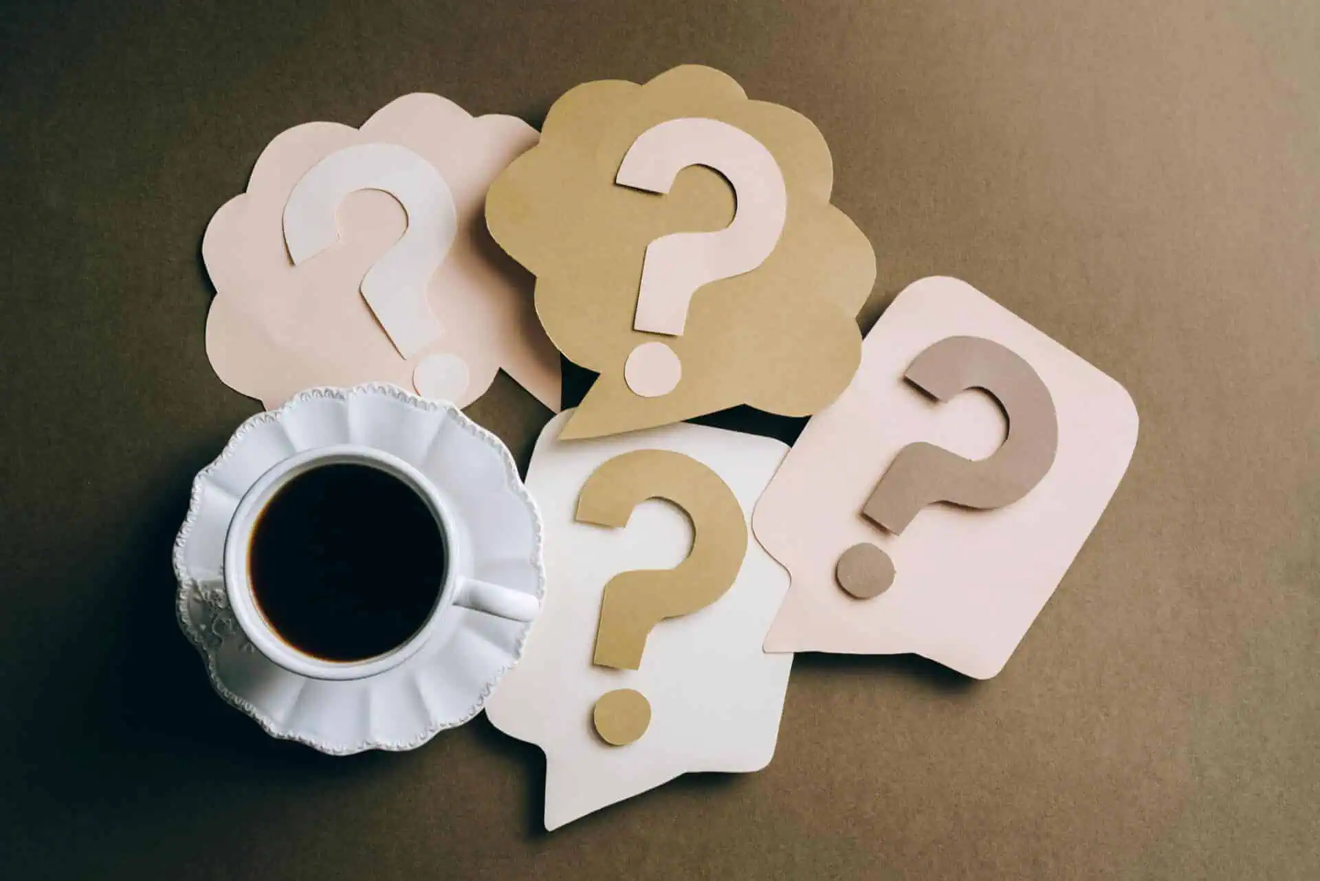Eine Tasse schwarzen Kaffees steht auf einer braunen Fläche neben Papierausschnitten in Form von Sprech- und Denkblasen, die jeweils mit einem großen Fragezeichen versehen sind - perfekt für ein Brainstorming von FAQ oder häufig gestellten Fragen.