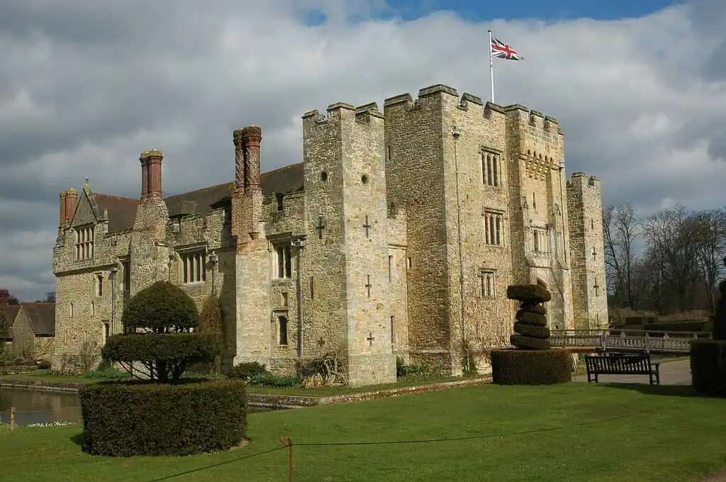 Ein historisches Schloss aus Stein mit Zinnen, hohen Schornsteinen und einer Union-Jack-Flagge auf der Spitze, umgeben von gestutzten Hecken, Gras und einem Graben unter einem bewölkten Himmel.