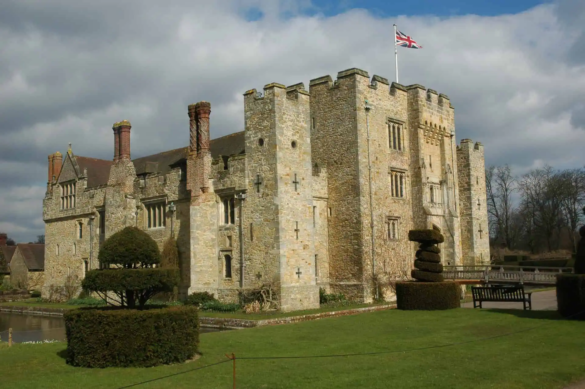 Ein historisches Schloss aus Stein mit Zinnen, hohen Schornsteinen und einer Union-Jack-Flagge auf der Spitze, umgeben von gestutzten Hecken, Gras und einem Graben unter einem bewölkten Himmel.
