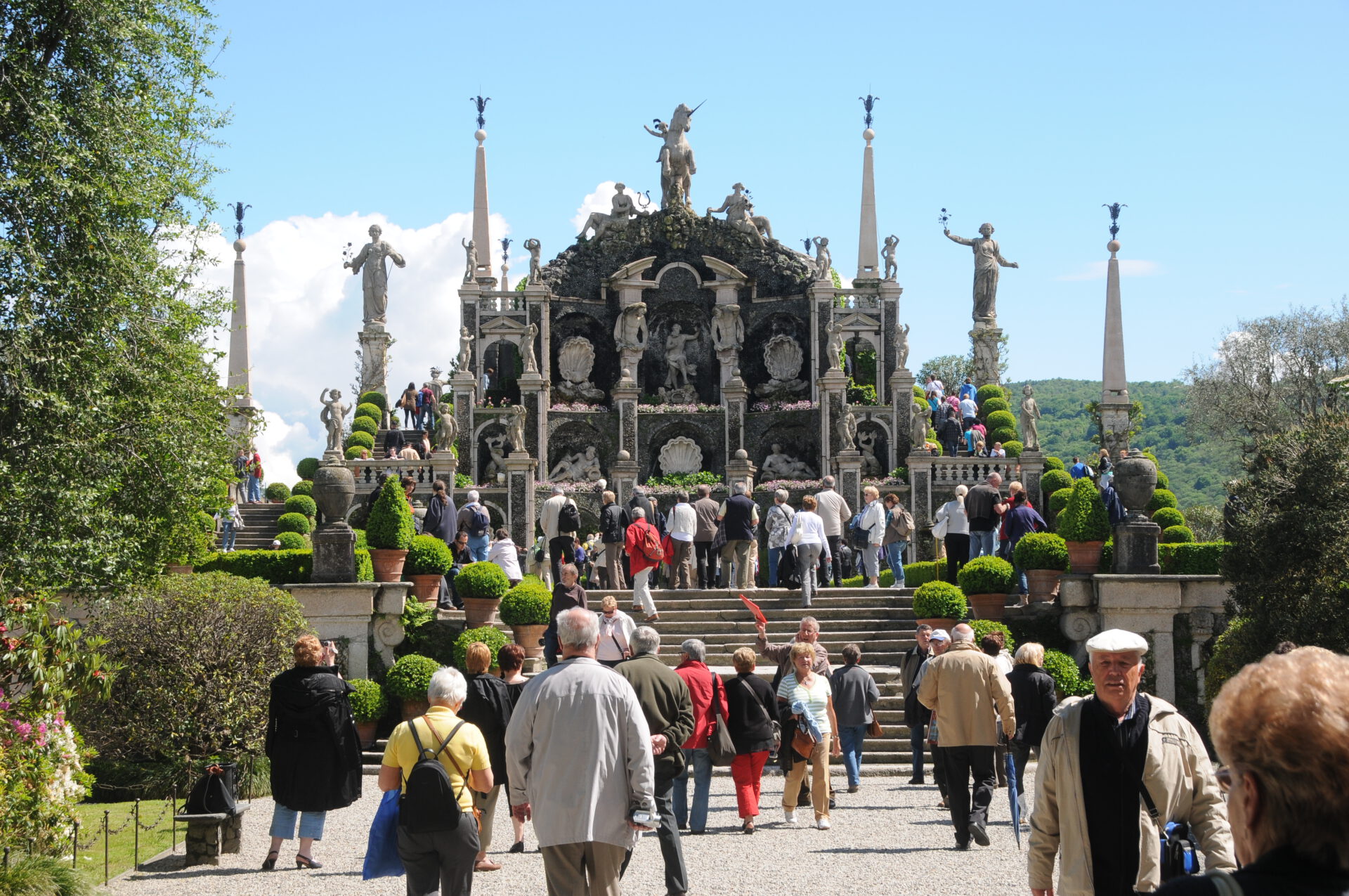 Eine Menschenmenge geht an einem sonnigen Tag auf eine große, verzierte Steinterrasse mit Statuen, Säulen und dekorativem Grün vor einer Kulisse aus Bäumen und fernen Hügeln zu.