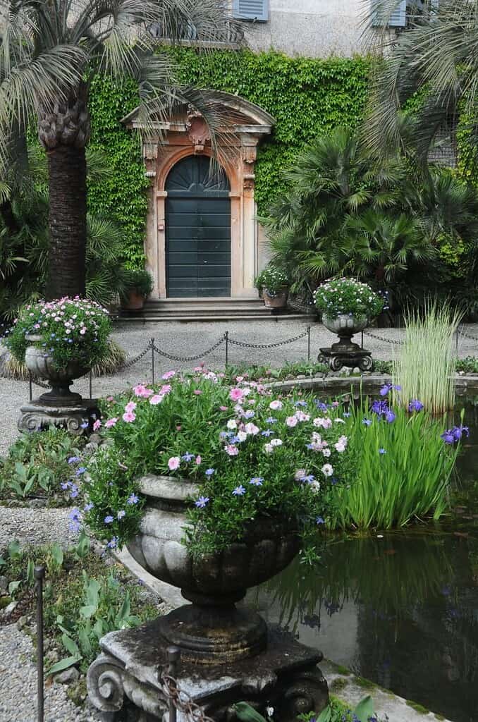 Große Steinkübel mit rosa, violetten und weißen Blumen stehen neben einem kleinen Teich in einem üppigen Garten, der an die ruhige Schönheit der Italienieschen Seen erinnert. Im Hintergrund ist ein hoher Torbogen zu sehen, der von efeubewachsenen Mauern und Palmen eingerahmt wird.