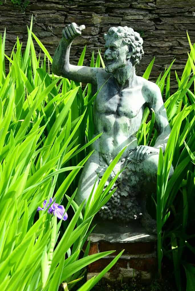 Eine Bronzestatue eines lächelnden, bärtigen Mannes sitzt inmitten hoher grüner Pflanzen an einer Steinmauer, mit einer einzelnen violetten Blume im Vordergrund - ein Hinweis auf den heiteren Charme der Italienieschen Seen.