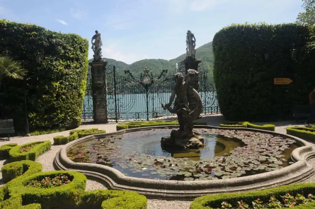 Ein dekorativer Brunnen mit einer Statue steht in einem Garten, der von gepflegten Hecken umgeben ist, mit einem verzierten Eisentor und Statuen, mit Blick auf einen malerischen See und grüne Hügel, die an die Italienieschen Seen erinnern.