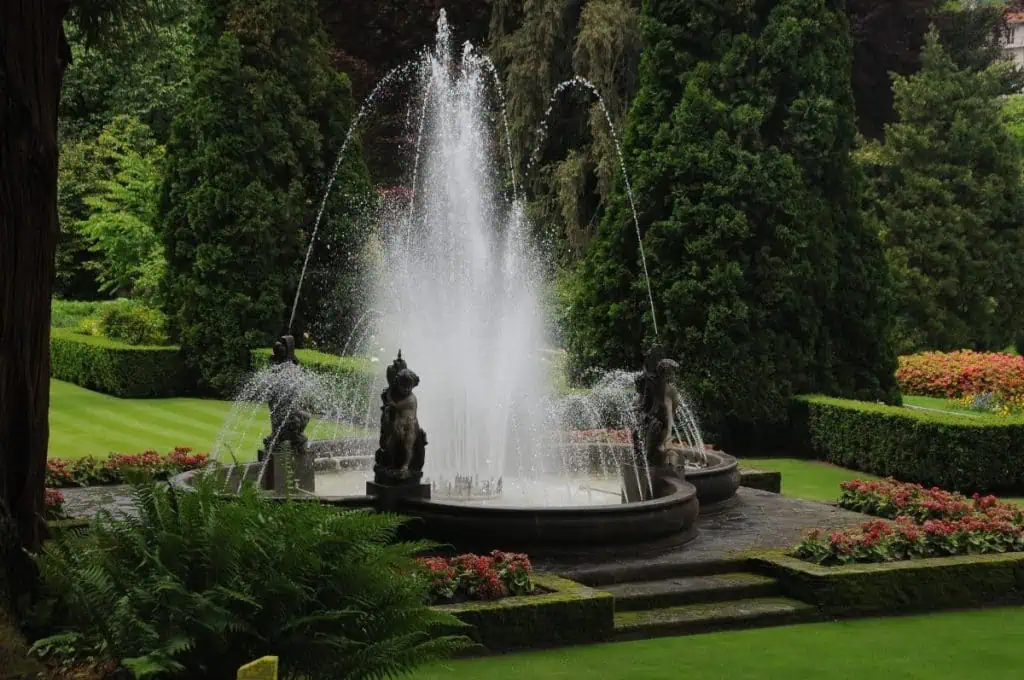 Ein kreisförmiger Steinbrunnen spritzt das Wasser hoch in die Luft, umgeben von üppigen grünen Rasenflächen, gepflegten Büschen und leuchtenden Blumen, die an die Eleganz der italienischen Seen erinnern. Statuen schmücken den Rand des Brunnens unter hohen Bäumen in einer Gartenlandschaft.