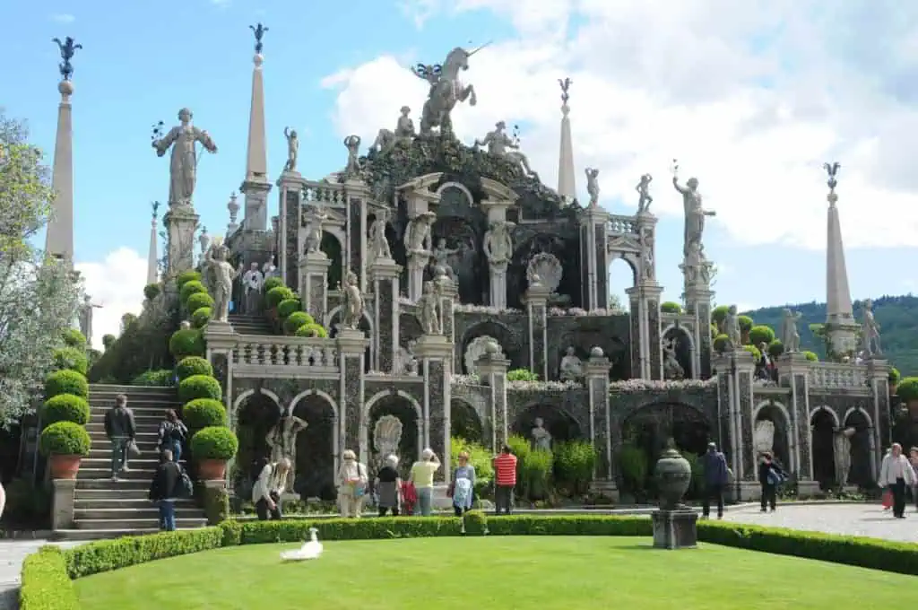 Eine große barocke Gartenterrasse mit kunstvollen Statuen, Bögen, üppigen grünen Hecken und Menschen, die flanieren und die Schönheit bewundern, erinnert an die Eleganz der Italienieschen Seen an einem hellen Tag.