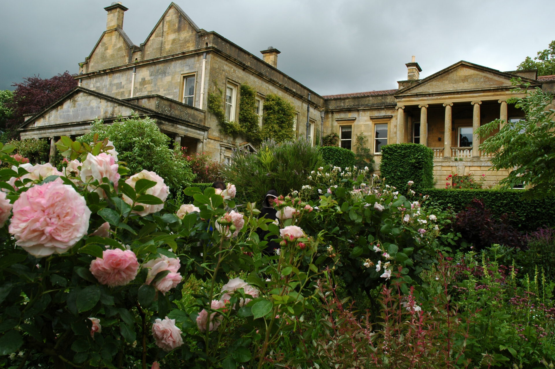 Ein großes Steinhaus mit Säulen und spitzen Dächern steht hinter einem üppigen Garten mit blühenden rosa Rosen und grünen Sträuchern unter einem bewölkten Himmel.
