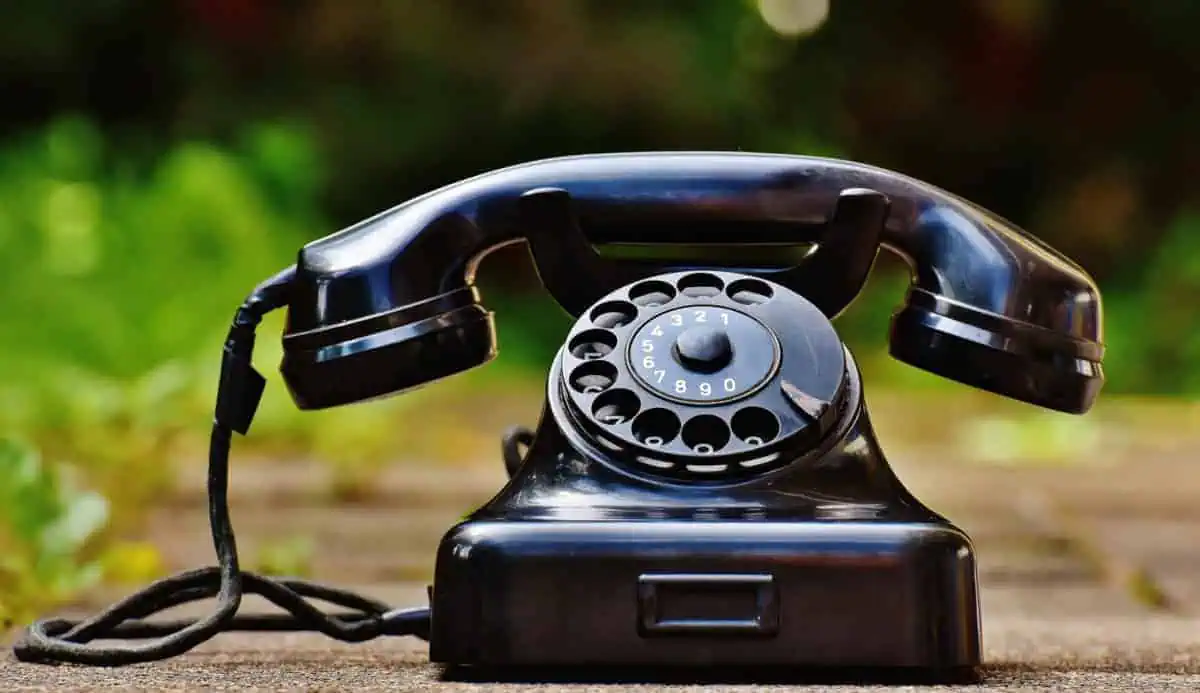 Ein altes schwarzes Wählscheibentelefon mit einer Schnur, das im Freien auf einer hölzernen Oberfläche mit einem unscharfen grünen Hintergrund steht.