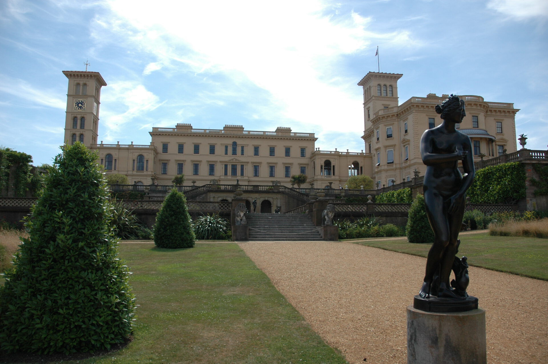 Ein großes, cremefarbenes Herrenhaus mit Türmen und vielen Fenstern steht hinter gepflegten Gärten und einem Kiesweg, auf dem im Vordergrund eine dunkle Frauenstatue steht. Der Himmel ist teilweise bewölkt.