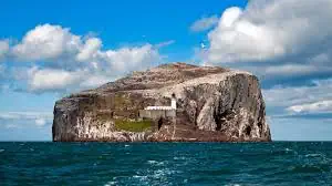 Eine große Felseninsel mit steilen Klippen erhebt sich aus dem blauen Ozean in Ost-Schottland. In der Nähe ihrer Basis steht ein kleines weißes Gebäude unter einem teilweise bewölkten Himmel.