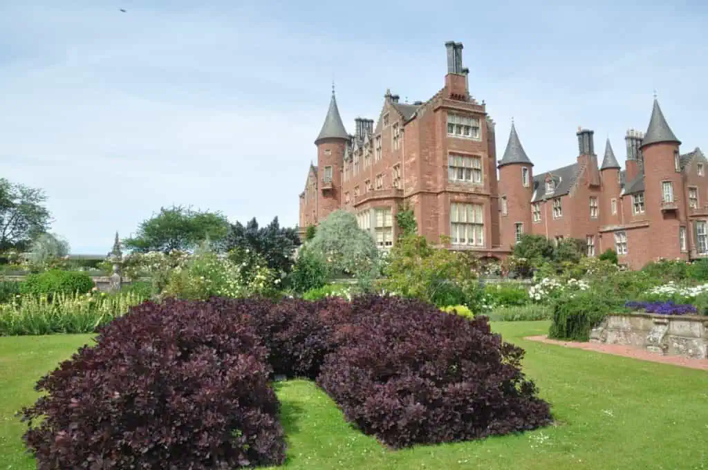 In Ost-Schottland steht ein großes Schloss aus rotem Backstein mit Türmchen, umgeben von üppigen Gärten, grünem Rasen, blühenden Sträuchern und ordentlich geschnittenen Hecken unter einem klaren Himmel.