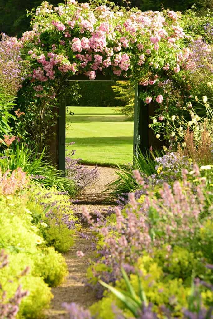 Ein Gartenweg, der an Ost-Schottland erinnert und mit üppigen grünen und violetten Pflanzen gesäumt ist, führt zu einem hölzernen Tor, das mit blühenden rosa Blumen bedeckt ist und auf einen gepflegten Rasen führt.