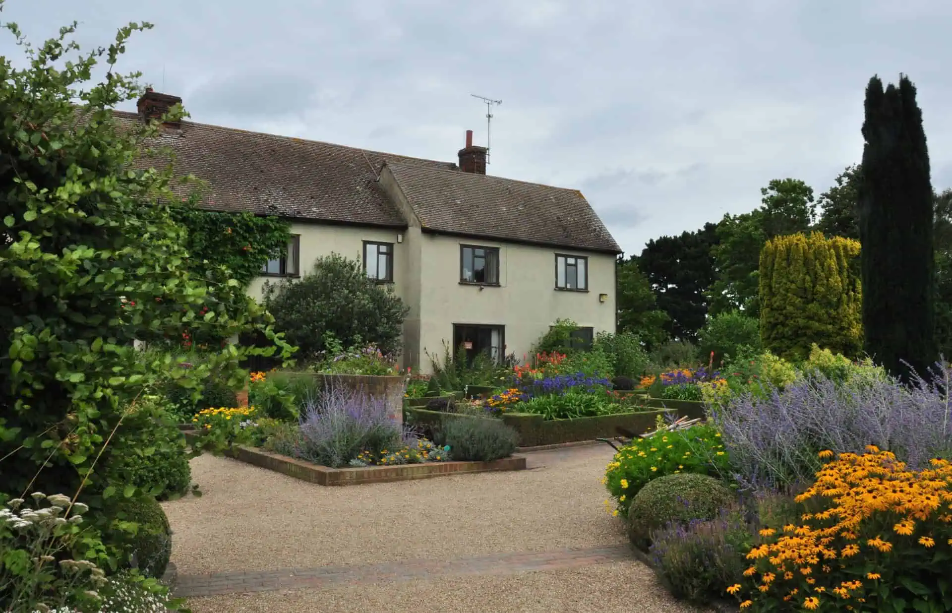 Ein zweistöckiges Haus mit beigem Äußeren ist von einem farbenfrohen, gut gepflegten Garten mit verschiedenen Blumen, Sträuchern und ordentlich gestutzten Büschen unter einem bedeckten Himmel umgeben.