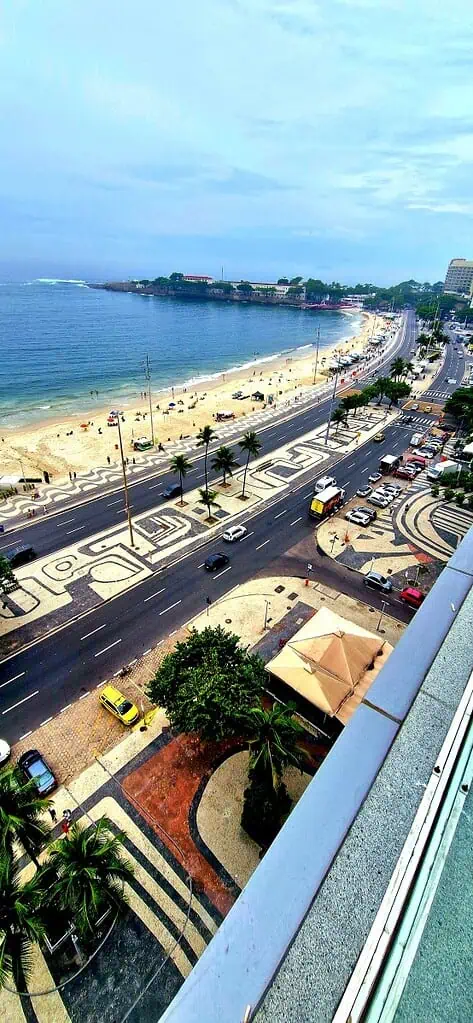 Luftaufnahme der Küstenlinie von Rio de Janeiro mit einem Strand, Palmen und einer breiten Straße voller Autos und Busse. Die Küstenlinie wölbt sich, während sich die Menschen im Sand entspannen und die Wellen unter einem teilweise bewölkten Himmel anrollen.