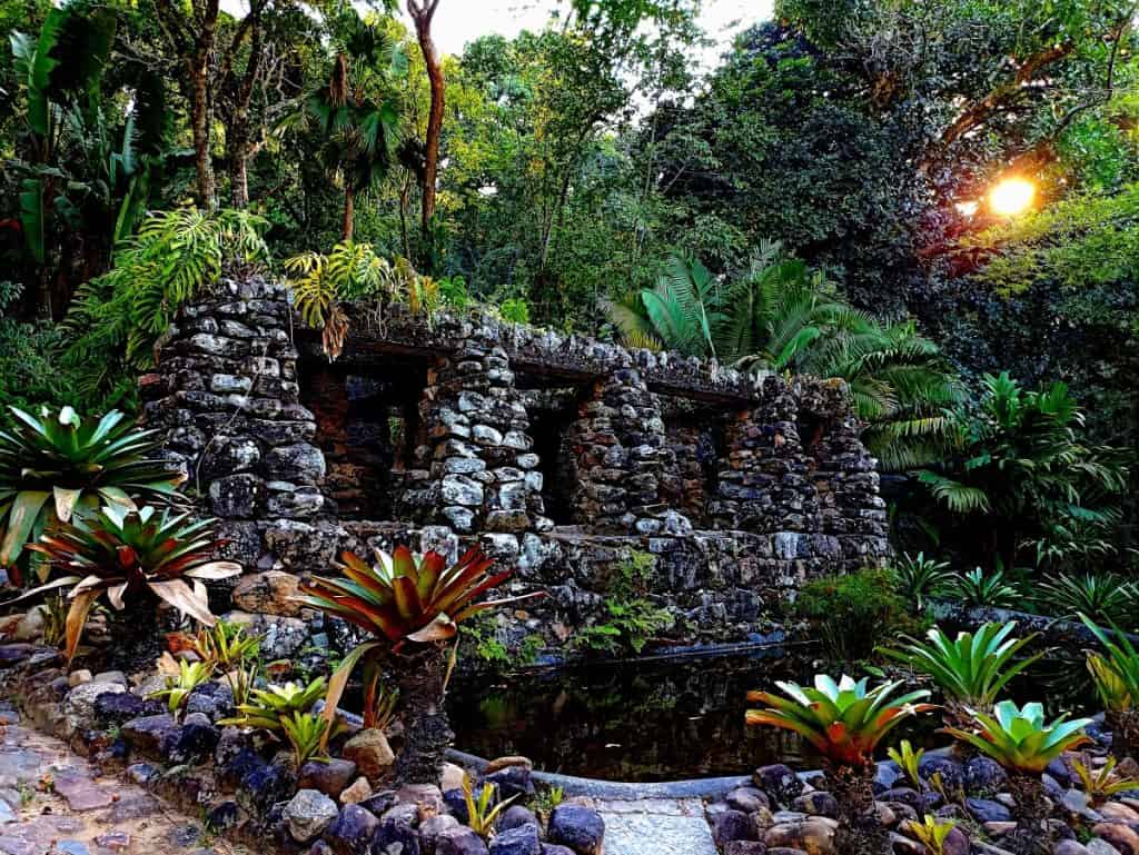 Ein altes Steinbauwerk mit drei Bögen ist von üppigen tropischen Pflanzen und Bäumen umgeben, und das Sonnenlicht, das durch den Wald im Hintergrund fällt, erinnert an den historischen Charme von Rio de Janeiro.