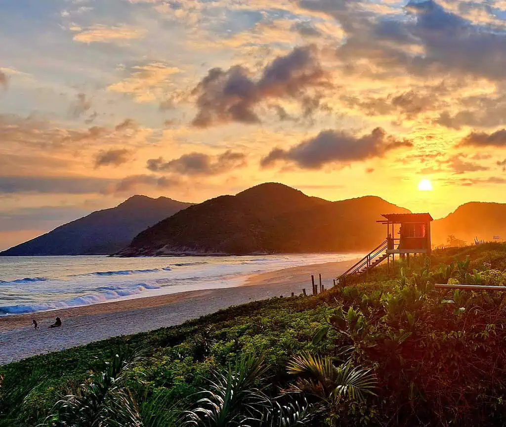 Ein farbenfroher Sonnenuntergang über einem Sandstrand in Rio de Janeiro mit sanften Wellen, fernen Bergen und einer Rettungsschwimmerhütte, die sich vom orangefarbenen Himmel abhebt. Üppiges Grün säumt den Vordergrund, während ein paar Menschen am Ufer entlang spazieren.