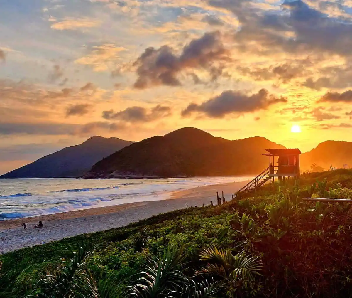 Ein farbenfroher Sonnenuntergang über einem Sandstrand in Rio de Janeiro mit sanften Wellen, fernen Bergen und einer Rettungsschwimmerhütte, die sich vom orangefarbenen Himmel abhebt. Üppiges Grün säumt den Vordergrund, während ein paar Menschen am Ufer entlang spazieren.