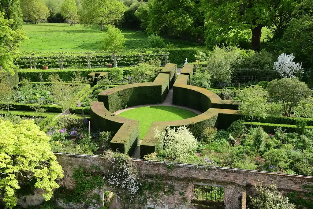 Ein formaler Garten mit einer kreisförmigen Rasenfläche, umgeben von ordentlich geschnittenen Hecken, geometrischen Wegen und üppigem Grün, an einem sonnigen Tag von oben betrachtet. Bäume und eine Ziegelmauer begrenzen den Garten.