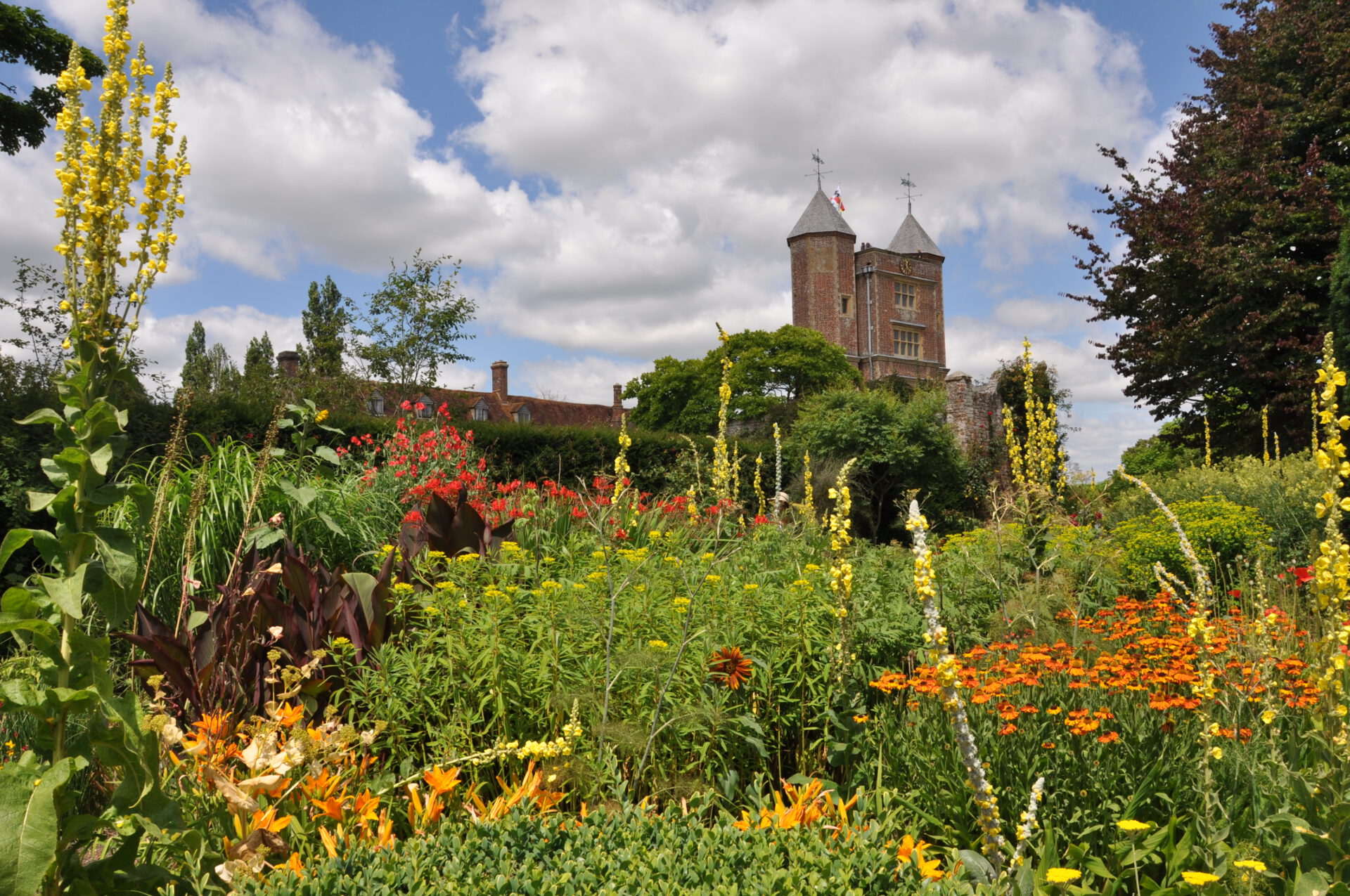 Farbenfroher Blumengarten mit gelben, orangefarbenen und roten Blüten im Vordergrund, üppige grüne Pflanzen und ein historischer Steinturm mit Türmchen im Hintergrund unter einem teilweise bewölkten Himmel.