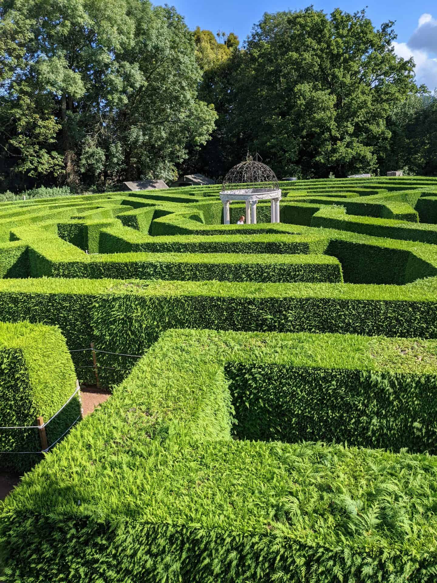 Ein üppiges grünes Heckenlabyrinth mit ordentlich gestutzten Mauern führt zu einem kleinen, offenen Pavillon mit einer dekorativen Metallkuppel, umgeben von hohen Bäumen unter einem teilweise bewölkten Himmel.