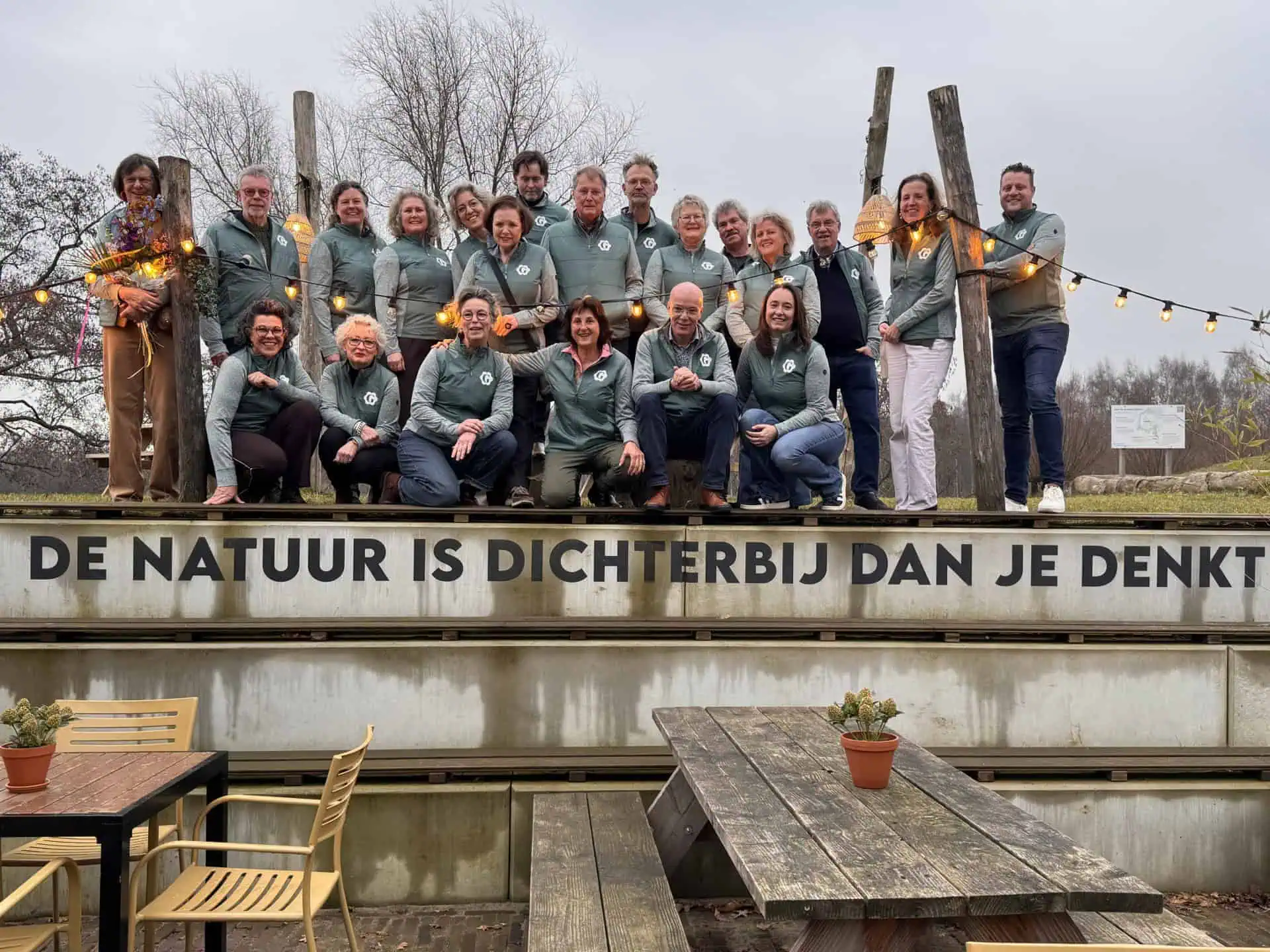 Sechs Erwachsene posieren im Freien in einer Gartenlandschaft. Drei sitzen auf einer beigen Couch, zwei stehen dahinter, und einer sitzt auf der Armlehne. Sie lächeln, umgeben von Grünzeug und hellem Tageslicht.