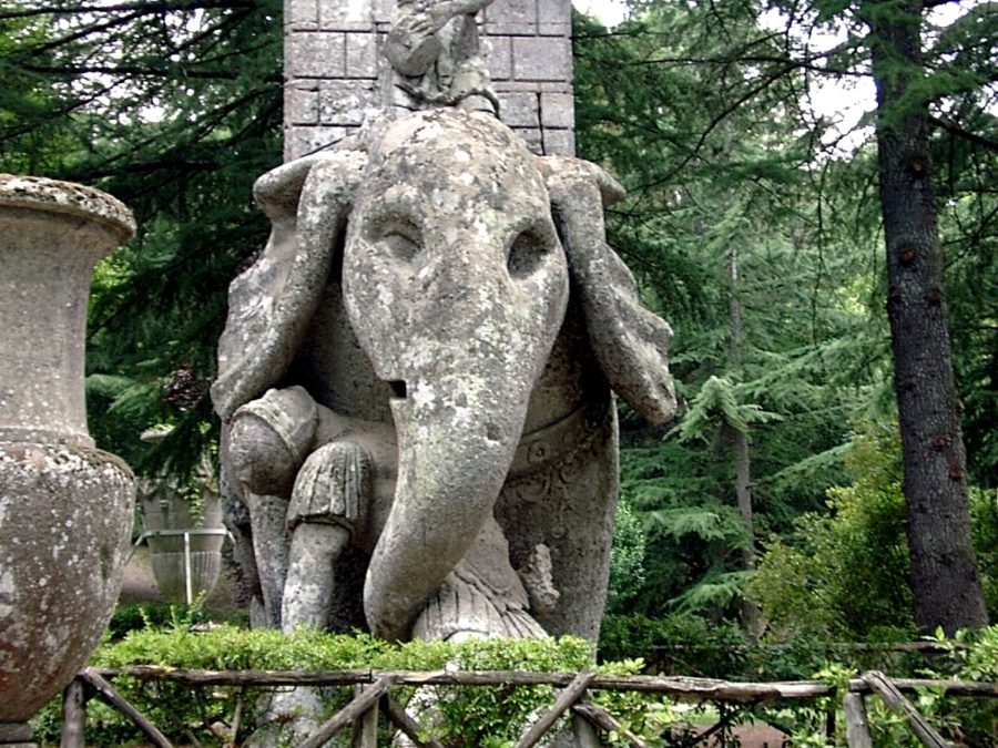 Eine steinerne Statue eines gepanzerten Elefanten steht in einem üppigen Garten, umgeben von Bäumen und Grün, mit einer Steinsäule und einem verzierten Zaun im Hintergrund.