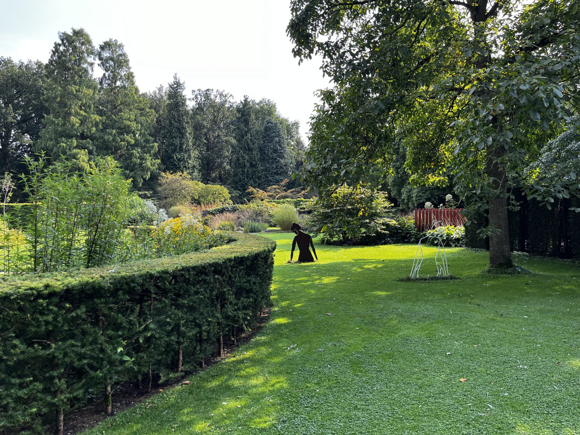 Ein üppiger grüner Garten mit gepflegten Hecken, hohen Bäumen und verschiedenen Sträuchern. Zwei abstrakte Skulpturen stehen auf dem Rasen, eine dunkel und sitzend, die andere weiß und kantig. Das Sonnenlicht fällt durch die Bäume.