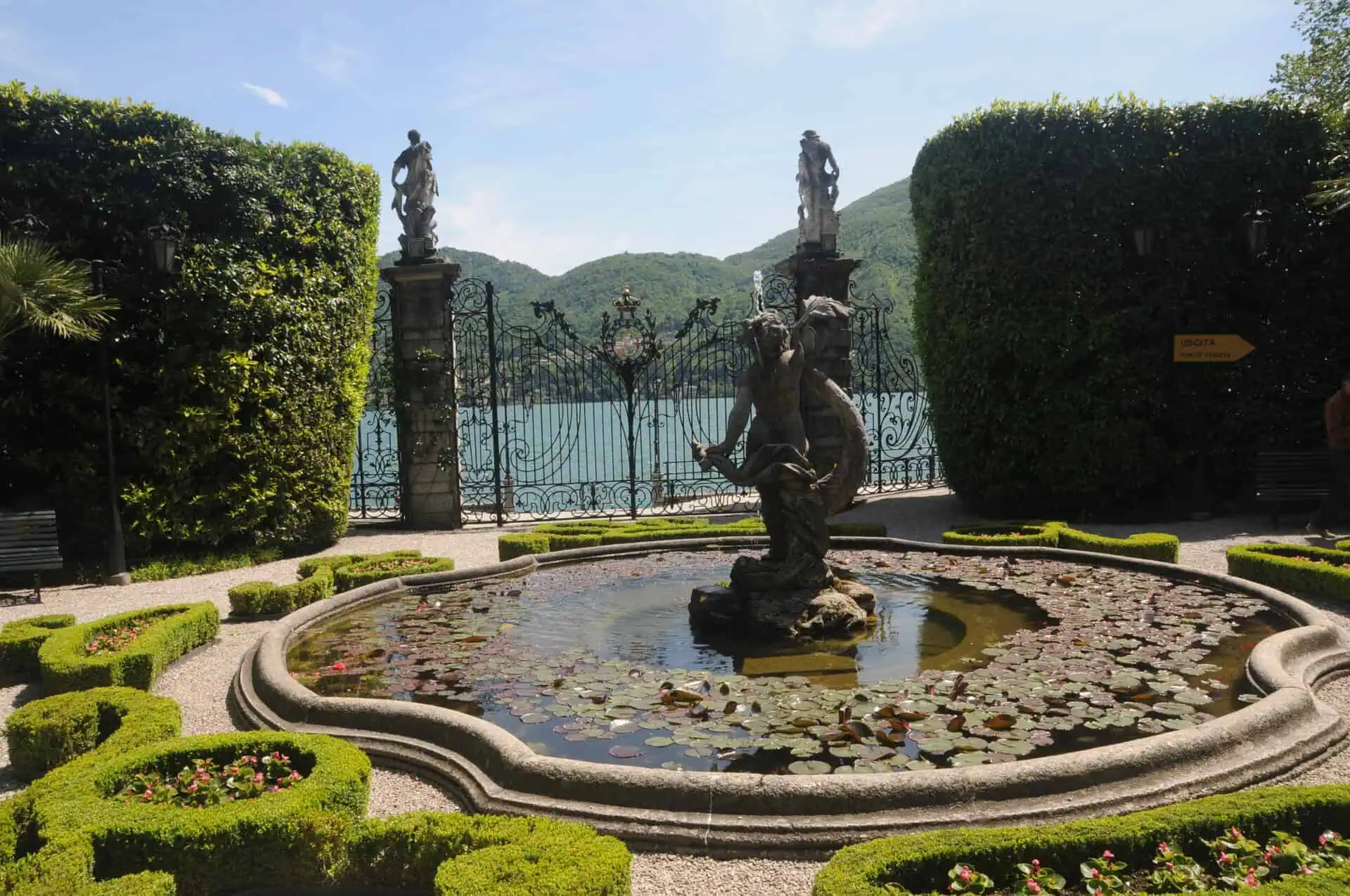 Ein dekorativer Springbrunnen mit einer Statue und Seerosenblättern ist von ordentlich geschnittenen Hecken und Blumen umgeben, mit verzierten Toren, Statuen, einem See und grünen Hügeln im Hintergrund unter einem blauen Himmel.